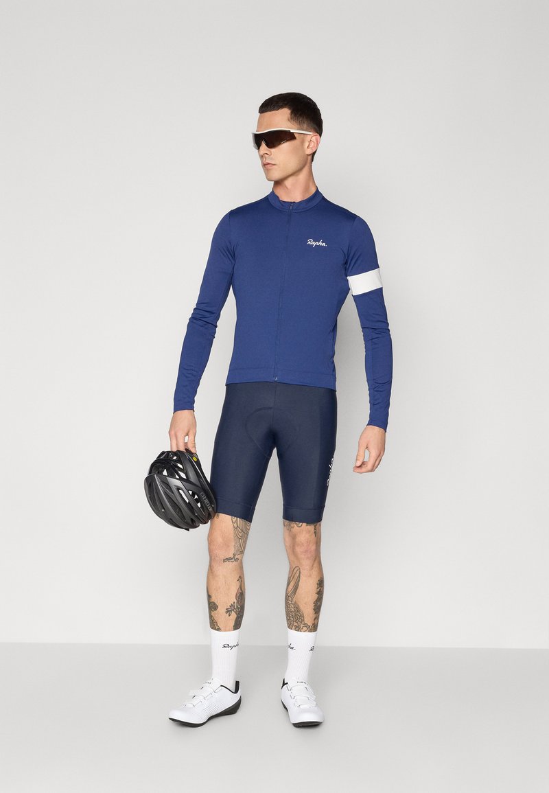 Marineblå langærmet cykeltrøje med en hvid stribe, parret med marineblå shorts. Modellen holder en sort hjelm og har solbriller samt hvide sko på.