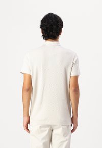 Polo à manches courtes beige clair avec un motif texturé, col classique et ourlet droit. Porté avec un pantalon de couleur claire.