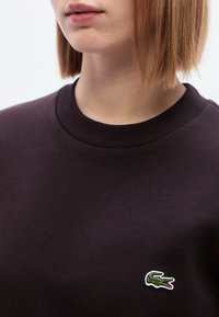 Σκούρο μωβ φούτερ με ριμπ ύφασμα στη στρογγυλή λαιμόκοψη και μικρό πράσινο λογότυπο κροκόδειλου στην αριστερή πλευρά του στήθους. Μαλακή υφή υφάσματος.