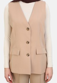 Gilet senza maniche beige con scollo a V, quattro bottoni e due tasche frontali, realizzato in un tessuto liscio, abbinato a una maglietta bianca a maniche lunghe.
