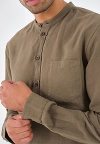 Camicia marrone con bottoni e colletto alla coreana, dotata di tasca sul petto e maniche lunghe in un tessuto strutturato. I bottoni sono visibili.