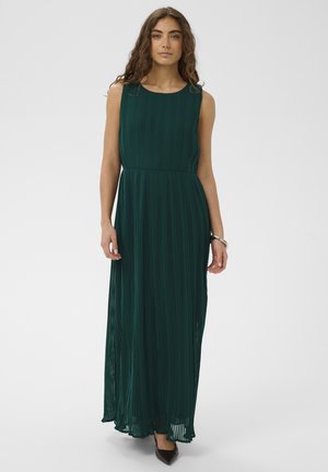 Mujer de pie con un vestido maxi plisado verde oscuro sin mangas y zapatos puntiagudos negros contra un fondo claro liso.