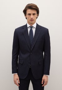 Marineblauw krijtstreeppak met reverskraag, twee knopen en zijzakken. Witte overhemd en een patroonstropdas maken de look af. Gladde textuur.