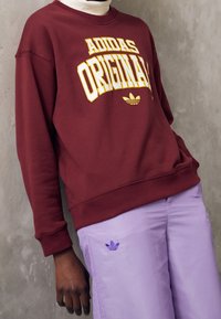 Mörkröd sweatshirt med "ADIDAS ORIGINAL" i gult, med rund halsringning. Stylen är kombinerad med ljuslila byxor som har en liten blå logotyp.
