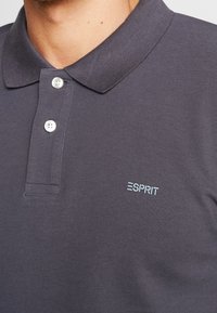 Polo shirt gris foncé en tissu texturé, avec un col classique, trois boutons blancs et un logo brodé bleu clair.