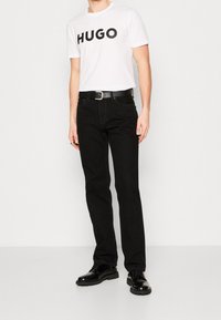 Mężczyzna w białej koszulce z napisem "HUGO" w boldzie, czarnych dżinsach, czarnym pasku i lśniących czarnych butach, stoi przy gładkiej ścianie.