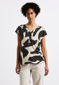 Top corto a maniche corte con motivo astratto in bianco e nero, con scollatura a V e dettaglio dei bottoni, realizzato in tessuto leggero, abbinato a pantaloni di colore chiaro.