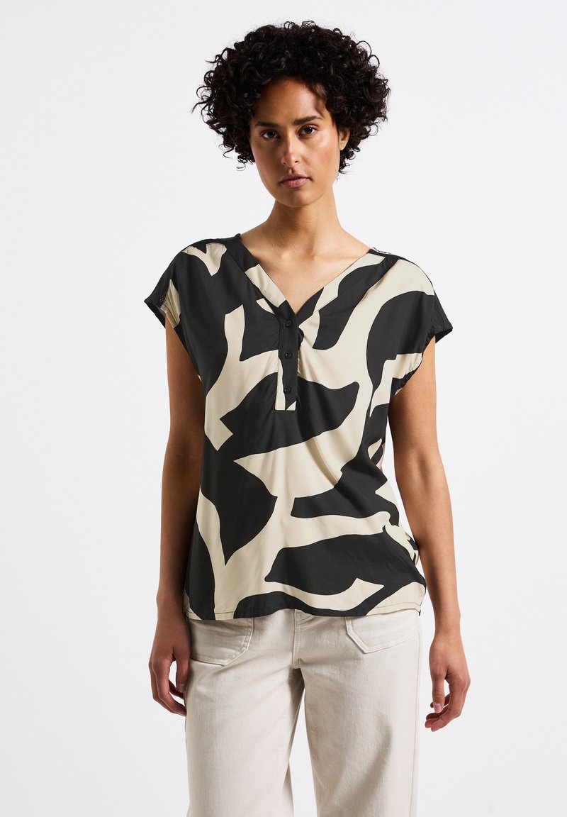 Top corto a maniche corte con motivo astratto in bianco e nero, con scollatura a V e dettaglio dei bottoni, realizzato in tessuto leggero, abbinato a pantaloni di colore chiaro.