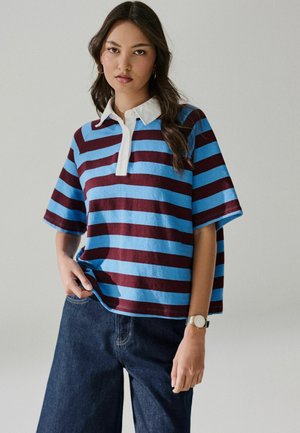Woman with long dark hair wearing blue and brown striped short-sleeve polo shirt and dark wide-leg jeans, standing against a plain background.