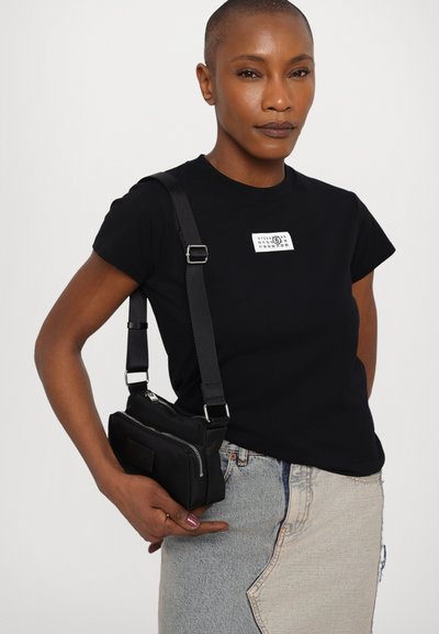 Black cotton t-shirt with short sleeves and a white label on the chest, paired with a small black crossbody bag featuring a zip closure.