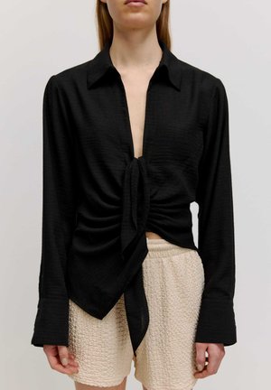 Zwarte, lange mouwen shirt met een diepe V-hals en knoopdetail in de taille. Textuurstof en gestructureerde kraag. Gecombineerd met gestructureerde beige shorts.