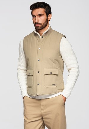 Uomo che indossa un gilet trapuntato beige con bottoni a pressione sopra un maglione bianco lavorato a trecce e pantaloni beige, in piedi con le mani nelle tasche.