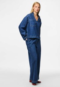 Denims stilizēts brīvajā laikā, kurā iekļauta brīvi pieguļoša, garām piedurknēm apģērbta virsma ar krūšu kabatu un plašas kājas bikses, abas tumši zilas krāsas.