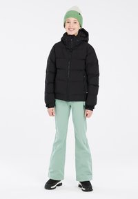 Giubbotto a giacca di piuma nera, con cappuccio e chiusura con zip; abbinato a pants da neve verde chiaro e stivali neri. Il modello indossa un berretto lavorato a maglia verde e crema.