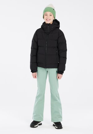 Schwarze Daunenjacke, mit Kapuze, mit Reißverschluss; kombiniert mit hellgrünen Schneehosen, schwarzen Stiefeln. Das Modell trägt eine grün-weiße Strickmütze.