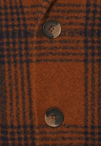 Tissu en mélange de laine marron avec un motif à carreaux bleu, comprenant un bouton noir à quatre trous. Texture douce, coupe sur mesure, et lignes épurées.