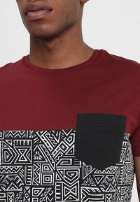 Rød t-shirt med en sort mønstret nederste halvdel og sort lomme. Materialet ser glat ud; mønstret indeholder geometriske former i hvid.