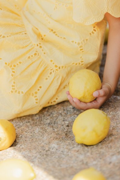 Rumena obleka z rožnatim vzorcem, ki drži teksturirano rumeno limono, obkrožena z dodatnimi limonami na kamniti površini.