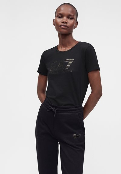 Жена, стояща с ръце зад гърба, облечена в черна тениска EA7 Emporio Armani и черен панталон в същия стил.