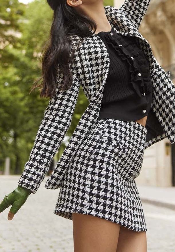 Veste à motif pied-de-poule noir et blanc et shorts assortis, avec un haut noir à volants en dessous et des gants verts ; textures lisses.