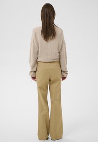 Femme aux longs cheveux bruns portant un pull en maille beige et un pantalon kaki à jambes larges, debout de dos sur un fond uni.