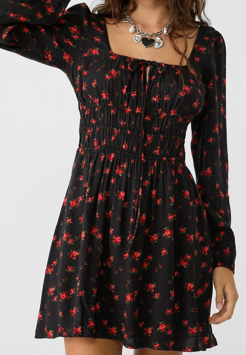 Femme portant une robe noire à manches longues avec imprimé floral rouge et taille froncée, associée à un collier chaîne argenté orné de charms en forme de cœur et de perles.