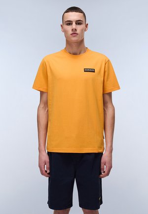 Gul bomulds t-shirt med korte ærmer, rund halsudskæring og et sort logo-patch på brystet. Stylet med mørke shorts.