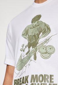 Bela bomba iz bombaža s zelenim grafičnim motivom mišičaste figure z utežmi, obdana z razpokanem tlem in besedilom "BREAK MORE SWEAT."