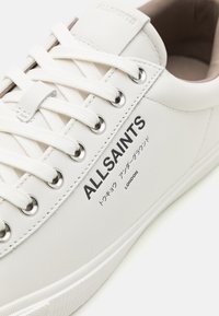 Adidași din piele albă cu vârf rotund, șase găuri pentru șireturi și șireturi plate. Prezentă branding imprimat și o talpă din cauciuc texturată.