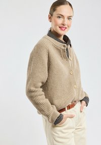 Beige geribde cardigan met een hoge kraag, houten knopen en lange mouwen, gelaagd over een donker geruit shirt, gecombineerd met lichte broeken.