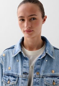 Giacca di denim azzurro chiaro con colletto classico, caratterizzata da decorazioni in tonalità oro e una finitura texturizzata, indossata su una camicia bianca.