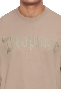Sudadera beige con un gran diseño de texto embosado y brillante que dice "Dropsize" en una fuente estilizada en el pecho.