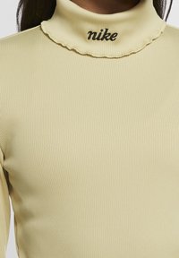 Ljusgul, ribbad turtleneck-top med en volangkrage och svart broderad "nike"-logotyp vid halsringningen. Mjuk, figurnära material.