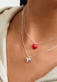Två lager halsband: ett med ett rött hjärta hänge, det andra med ett glänsande silverhjärta. Båda har smala, silverkedjor.