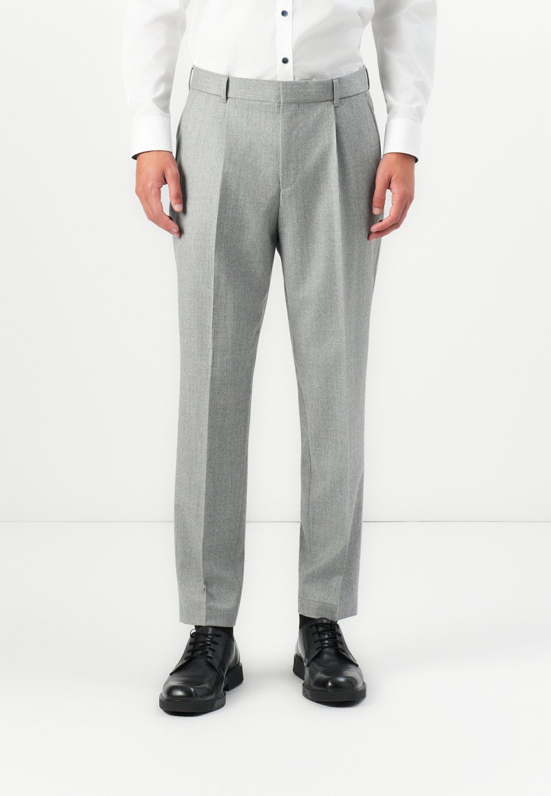 Man wearing light gray tailored trousers, black dress shoes, and a white buttoned shirt standing against a white background.
