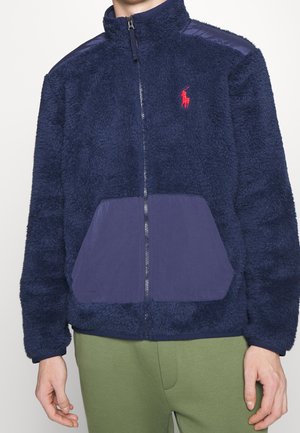 Veste polaire zippée bleu marine avec tissu texturé, poches avant lisses, logo rouge brodé sur la poitrine, portée avec un pantalon vert.