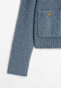 Cardigan en tricot texturé bleu avec une poche avant et un bouton en or. Présente des détails de poignet côtelés et un aspect de tissu doux.
