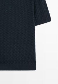 Chemise en maille bleu marine foncé à manches courtes avec motif tressé texturé et ourlet et bords de manches côtelés sur fond blanc.