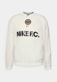 Biała kurtka z nylonu, z okrągłym dekoltem, elastycznymi mankietami, wypisanym wyraźnymi czarnymi literami "Nike F.C." oraz okrągłym detalem z logo.