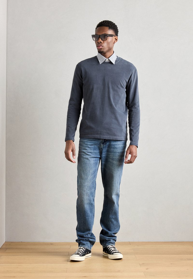 Graues Langarmshirt über einem weißen Hemd mit Kragen, blaue Jeans und schwarze Sneaker mit weißen Sohlen. Brille und neutraler Hintergrund.