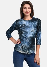 Blue floral print long-sleeve top with a textured surface, featuring a round neckline and a fitted design, paired with dark jeans.