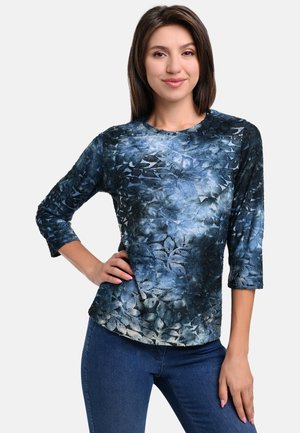Langärmliges Oberteil mit blauem Blumenmuster und strukturierter Oberfläche, mit Rundhalsausschnitt und taillierter Passform, kombiniert mit dunklen Jeans.