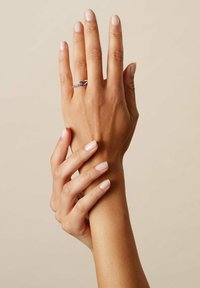 Silberner Ring mit gedrehtem Band, versehen mit einem kleinen, klaren Edelstein. Die Hände sanft positioniert, zeigen kurze, natürliche Nägel. Helle Hautfarbe.