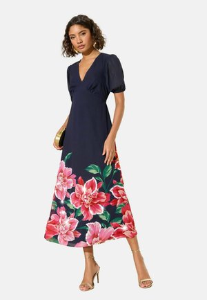 Žena v námornícky modrých midi šatách s nafúknutými rukávmi a veľkým ružovým kvetinovým vzorom na leme, drží zlatú kabelku, má na nohách zlaté lodičky.