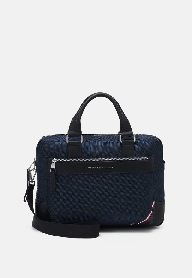Sacoche en tissu bleu marine avec des accents en cuir noir, deux poignées supérieures, une sangle réglable et une poche zippée à l'avant. Contient un détail du logo de la marque.