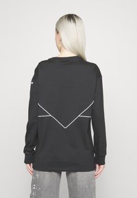 Person with long platinum blonde hair, wearing a black long-sleeve shirt with white geometric lines on the back and gray paint-splattered pants.