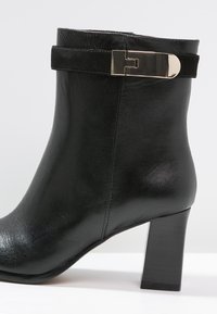 Botín negro de cuero con textura lisa, tacón grueso alto y una correa decorativa con un detalle de hebilla metálica.