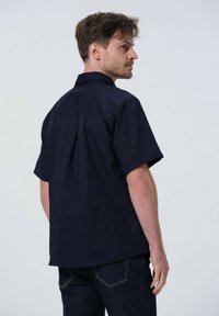 Chemise à manches courtes marine en tissu léger, avec un col pointu, un pli au dos et une coupe décontractée. Portée avec un jean en denim foncé.