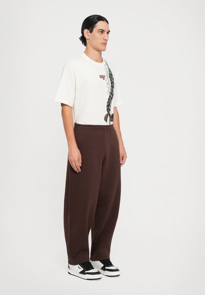 Weißes Baumwoll-T-Shirt mit Grafikdesign, kombiniert mit braunen Sweatpants. Das Modell trägt schwarz-weiße Sneaker; lässige Passform und elastischer Bund.