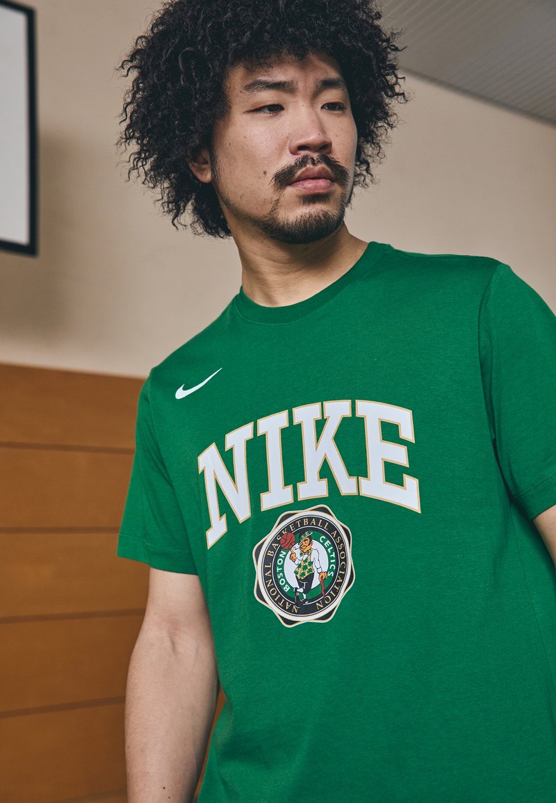 Grøn bomulds T-shirt med "NIKE" i hvid med en grå kant og et Boston Celtics-logo nederst til venstre. Rund hals design.